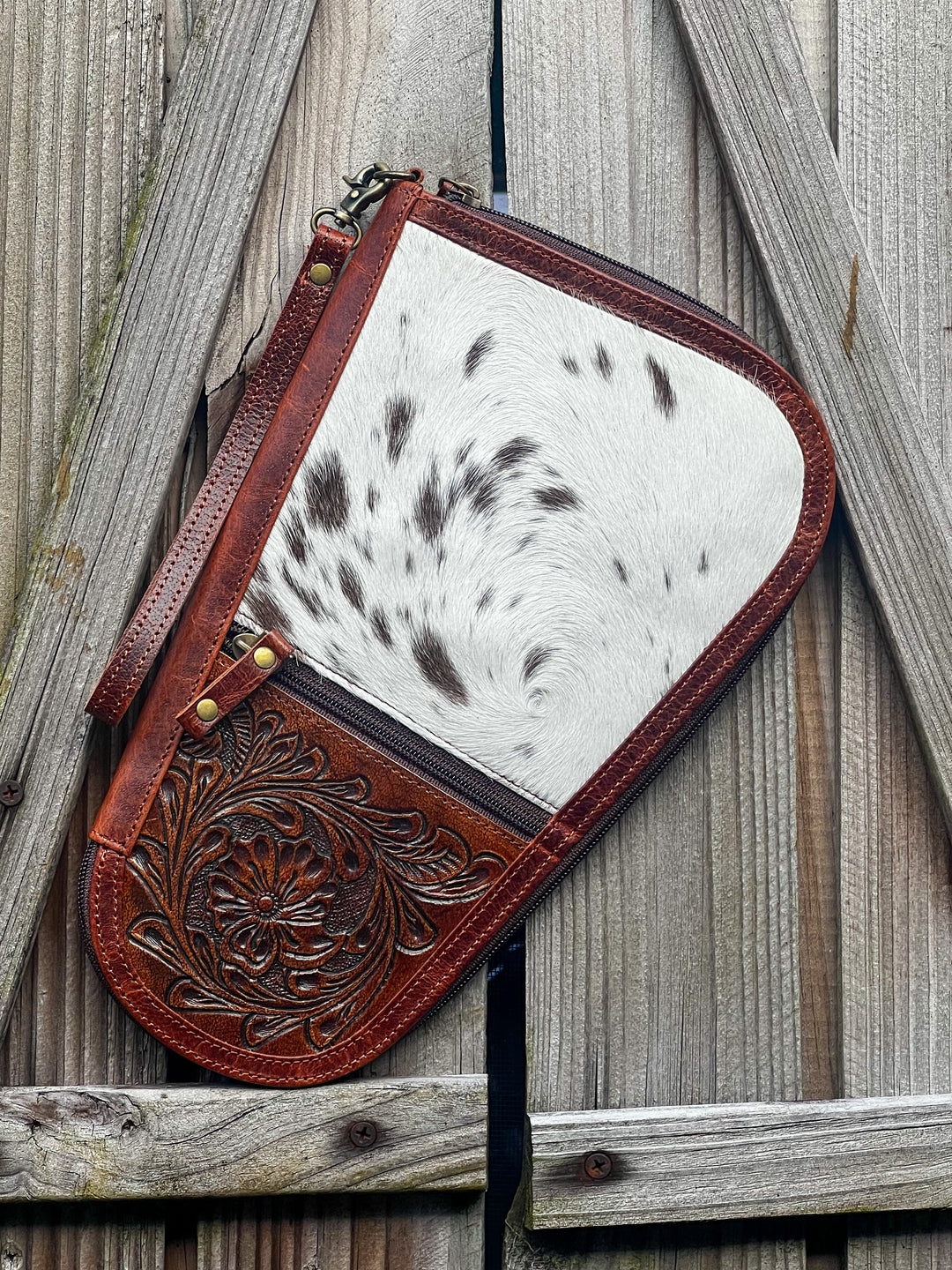 A handmade tooled leather and cowhide hand gun case with a front zipper, displayed against a wooden backdrop.