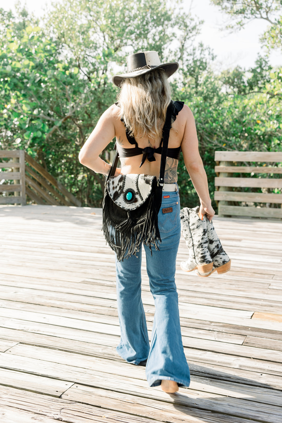 Woman walking barefoot in a coastal-western outfit carrying a black-and-white cowhide fringe crossbody bag and matching cowhide boots and hat.