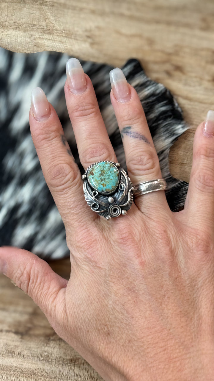 A close-up of a hand wearing a large Navajo handmade Sterling silver signed ring made my Jimmy Lee with a round mine 8 turquoise stone, set against a rustic wood and black-and-white cowhide background.