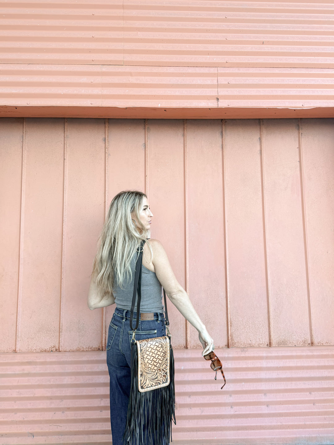 Woman with a tan and black tooled leather fringed crossbody purse against a pink wooden wall showing the size 