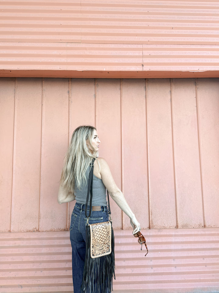 Woman with a tan and black tooled leather fringed crossbody purse against a pink wooden wall showing the size 