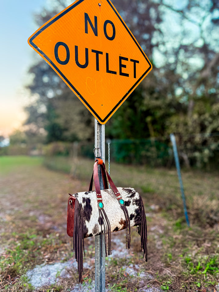 Cowhide leather duffel bag with fringe hanging from a 'No Outlet' sign in an outdoor setting.