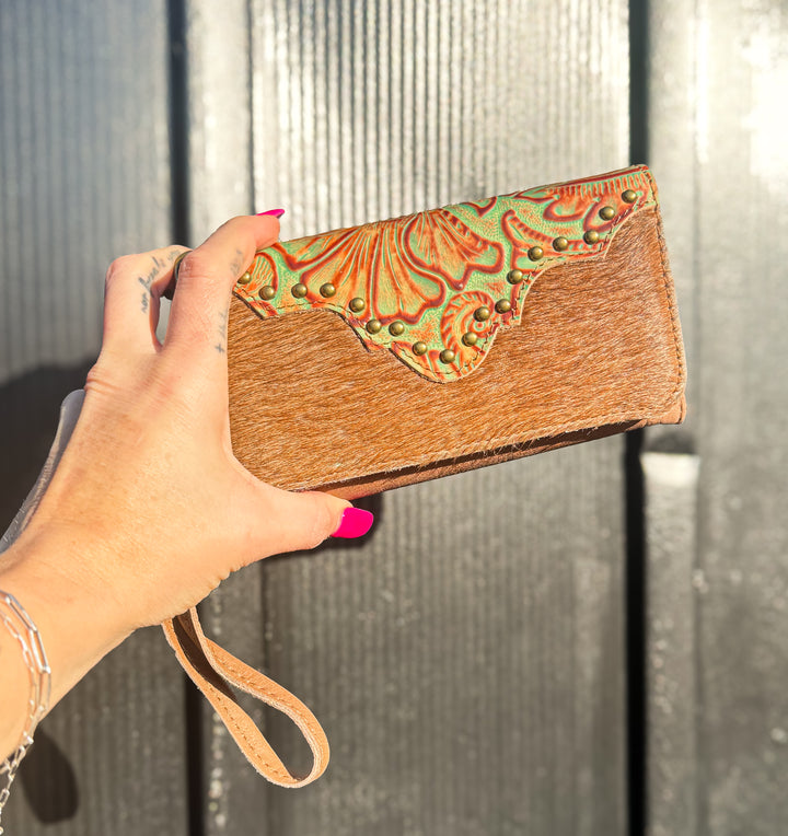 Hand holding a brown cowhide wallet with decorative floral embossed leather flap and hand strap against a dark background
