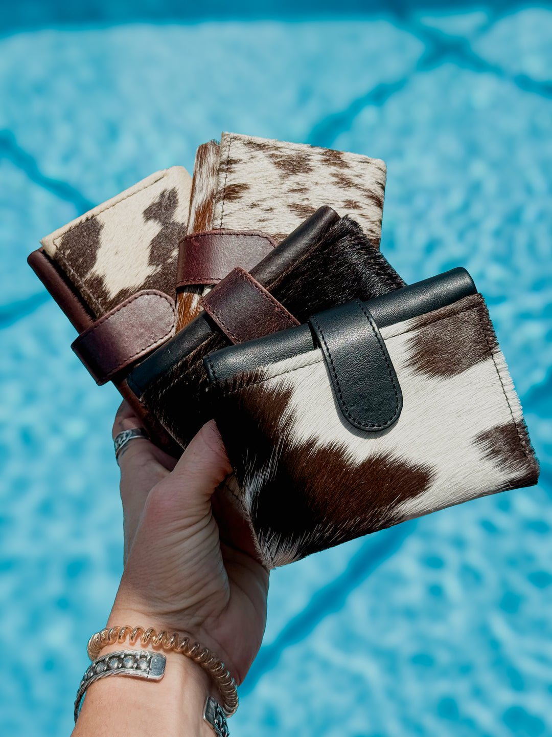 A hand holds four small trifold wallets with hair on cowhide and dark brown or black leather straps, set against a bright blue swimming pool background.