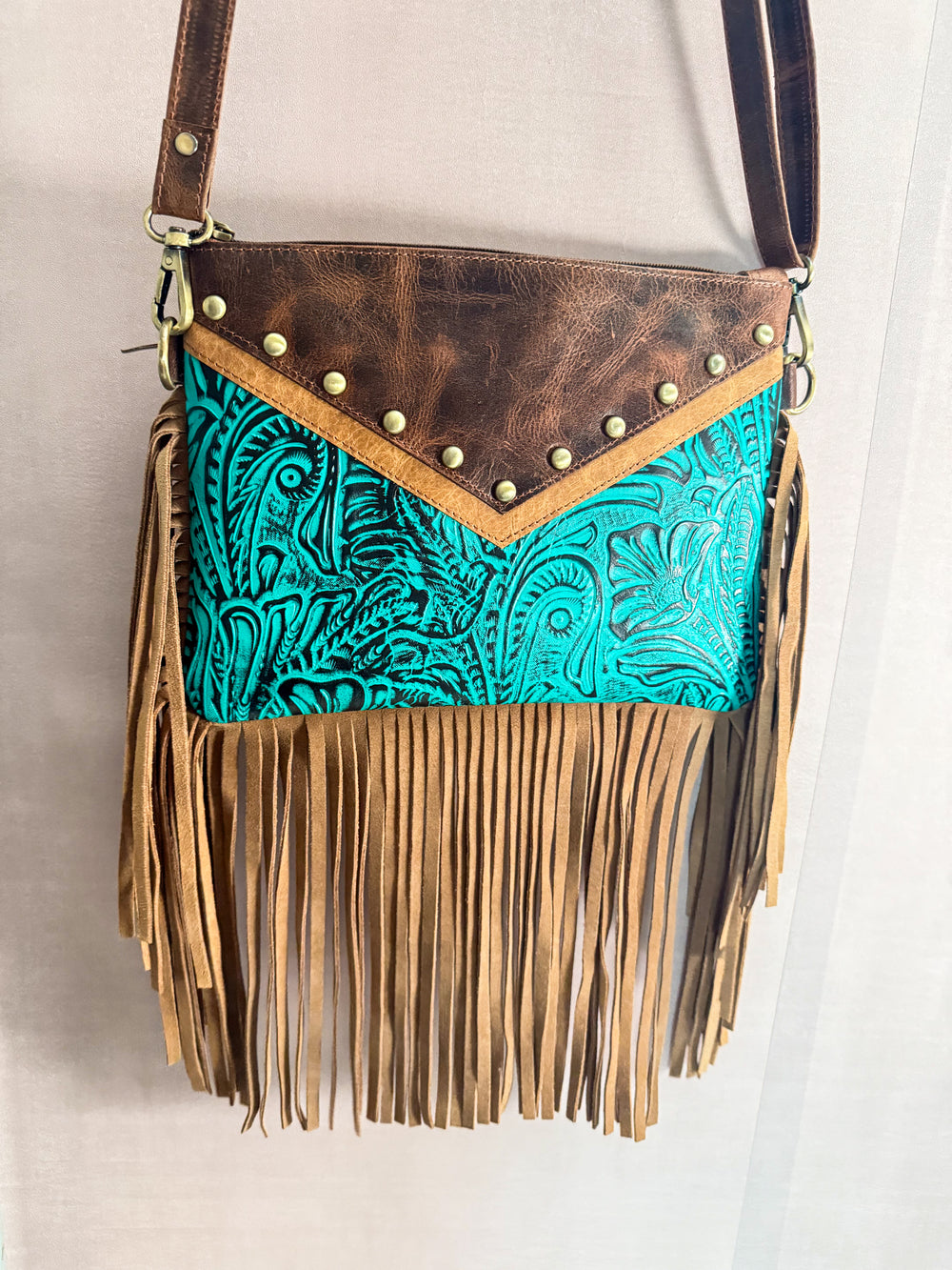 Brown leather crossbody bag with turquoise tooled leather and fringe on a light background