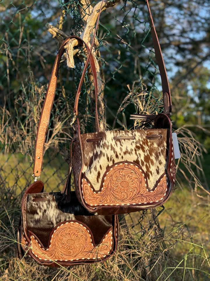Western cowhide bucket bag with hand-tooled leather detailing and adjustable strap, shown outdoors with natural rustic background