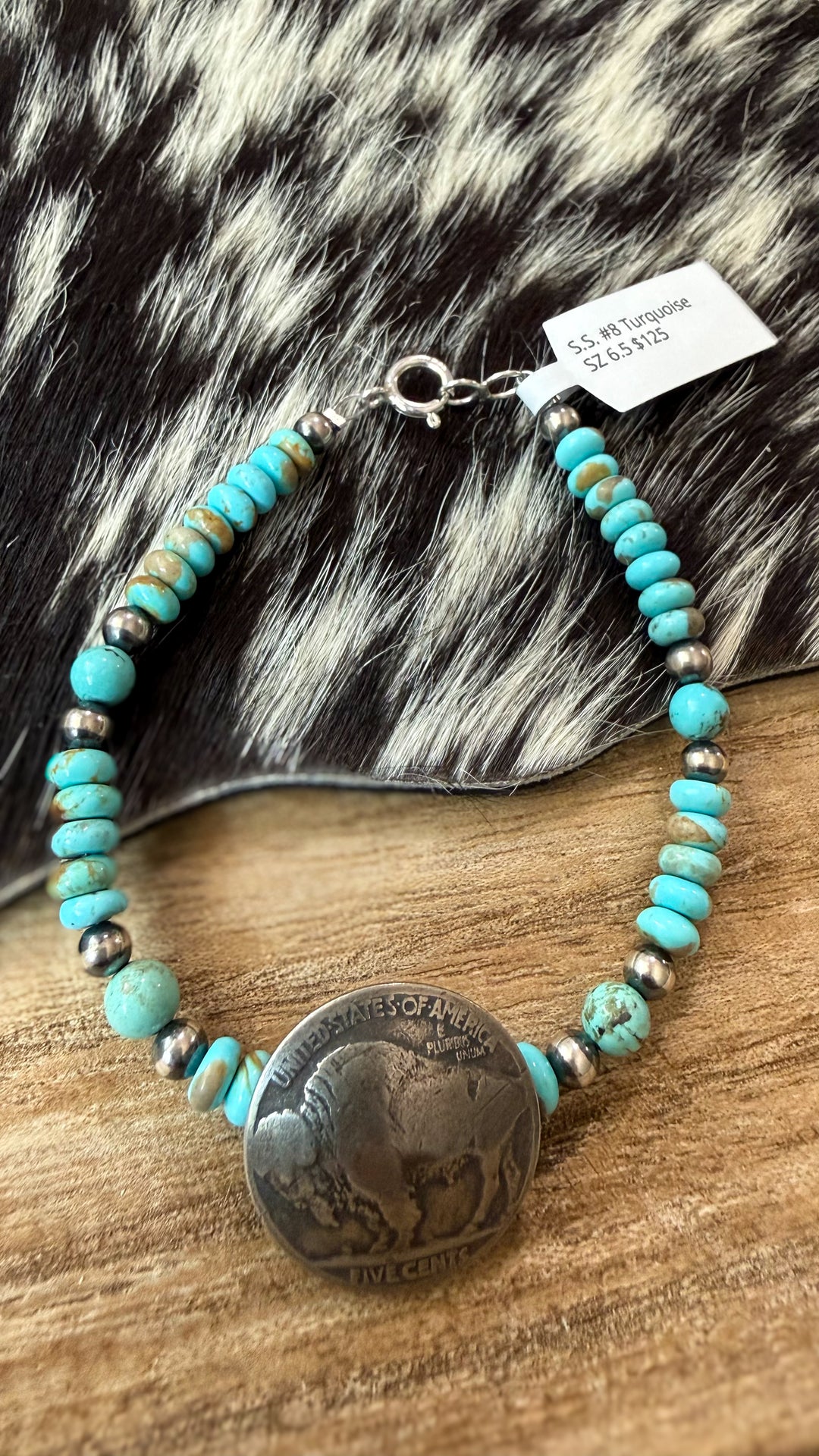 A Navajo handmade mine 8 Turquoise beaded bracelet with sterling silver Navajo pearl spacer beads and an authentic buffalo coin pendant center handmade by Navajo artist Kimberly Tom and shown on a wooden table with a cowhide coaster display