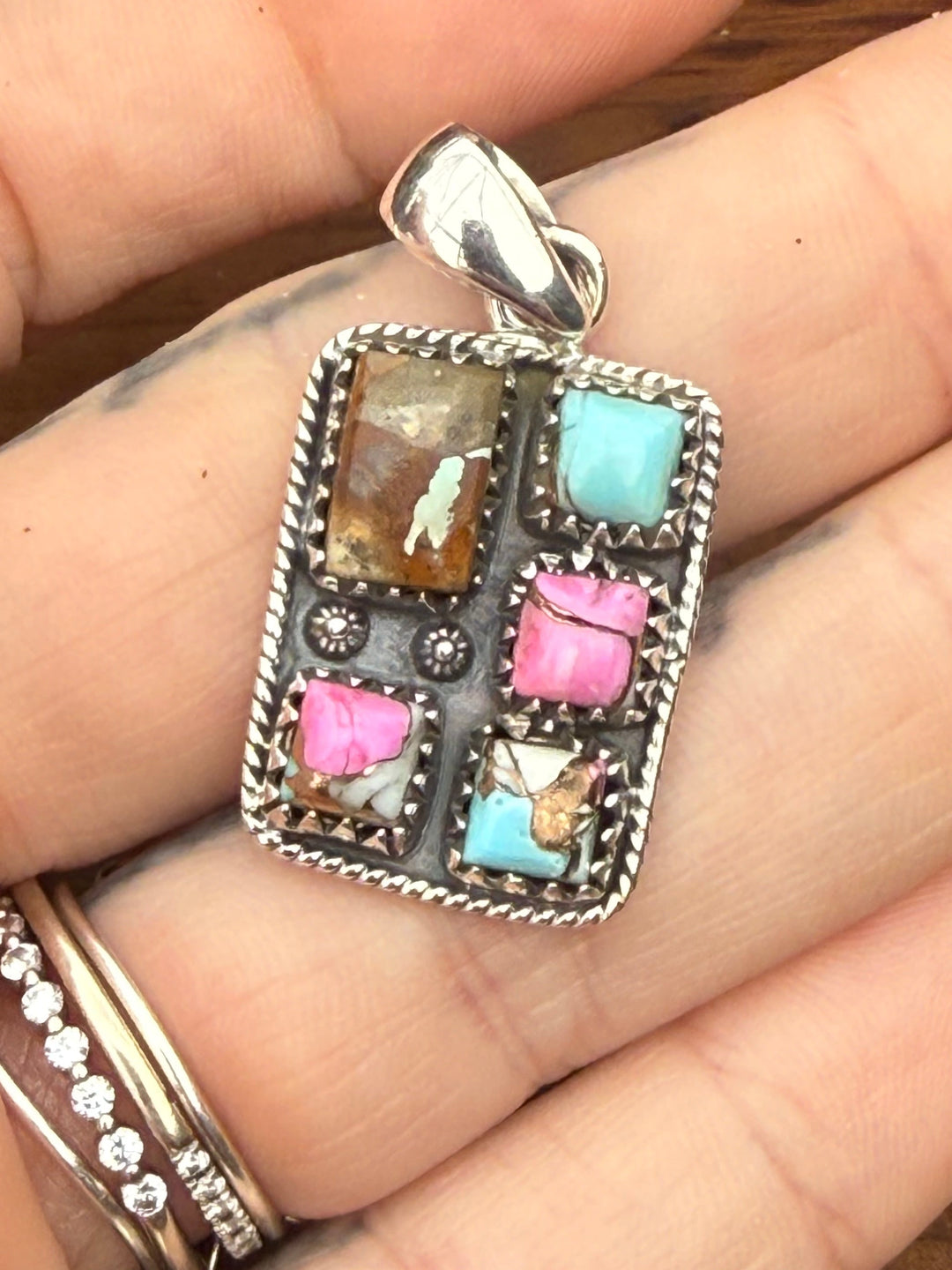 Hand holding Sterling silver Mine #8 turquoise and pink composite southwestern pendant with stamped detailing and bale on wood background