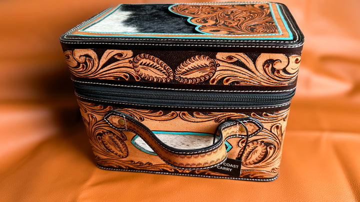 A rectangular zippered case with a tooled leather western design, featuring a black and white hair-on-hide top panel, turquoise accents, and a leather handle, set against a draped orange background by East Coast Carry