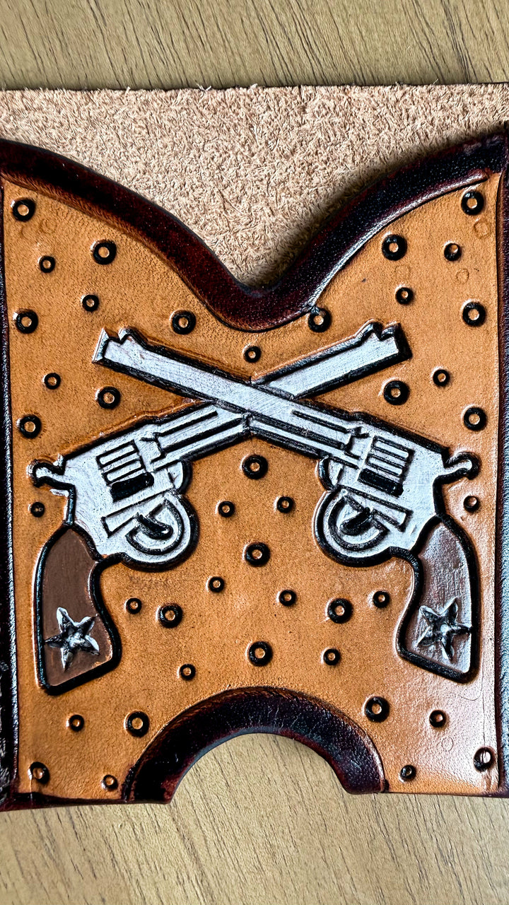 A close-up of a brown leather wallet or card holder featuring a tooled design of two crossed revolvers with silver-toned details and dark brown border accents, shown on a wood surface.