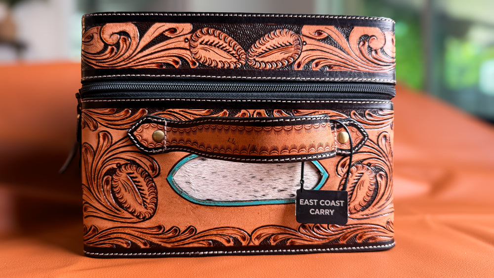 A tooled leather zippered case with ornate brown and black floral carving, a front leather handle, and a hair on cowhide white speckled deont panel. A black tag on the handle reads 'EAST COAST CARRY'.