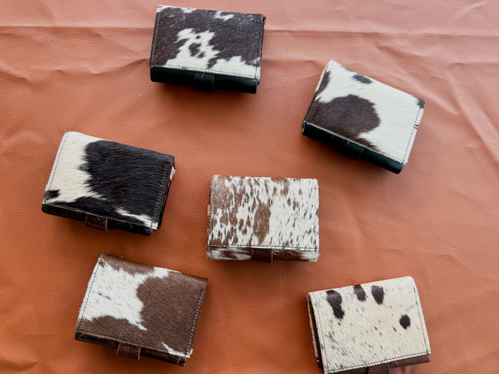 Back side of Six cowhide trifold wallets in black-and-white and brown-and-white natural hide patterns, arranged on a textured terracotta background.