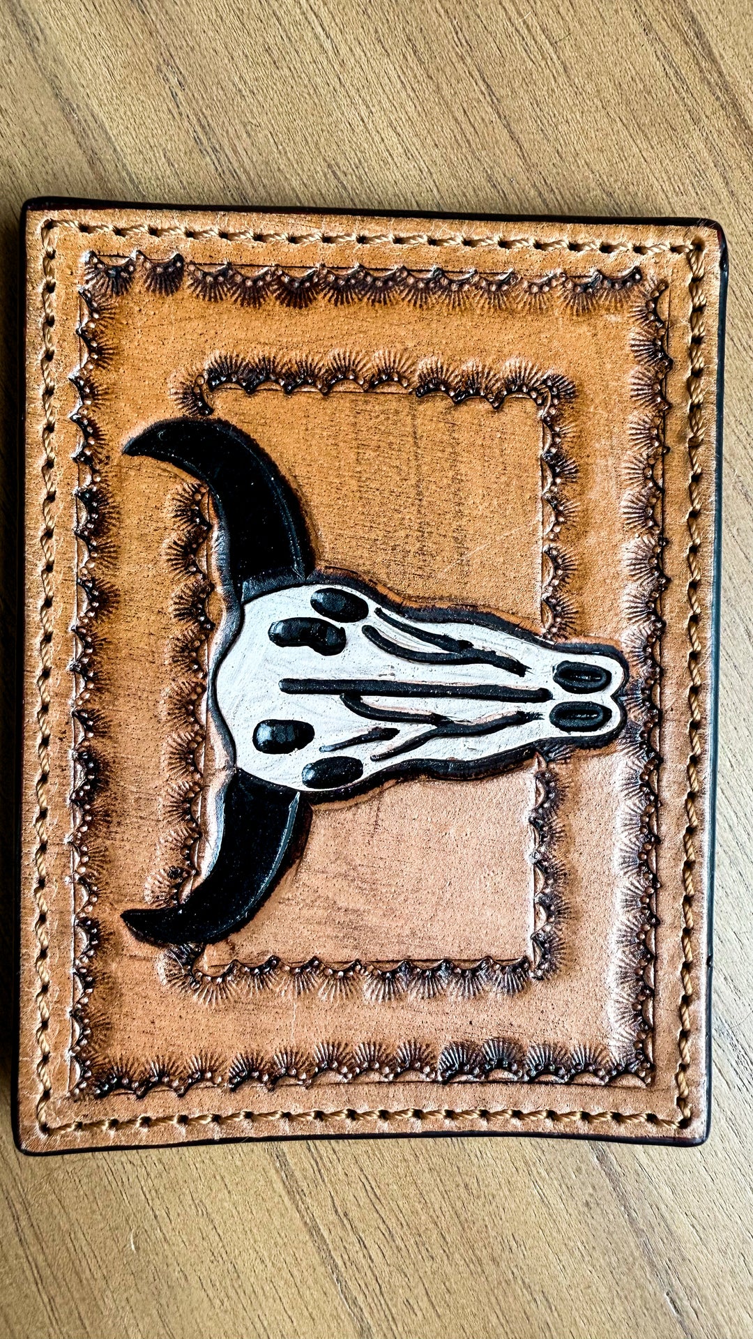 A tan leather wallet or card holder featuring a tooled border and a central cow skull patch with black horns and a white skull design, shown on a wooden surface.