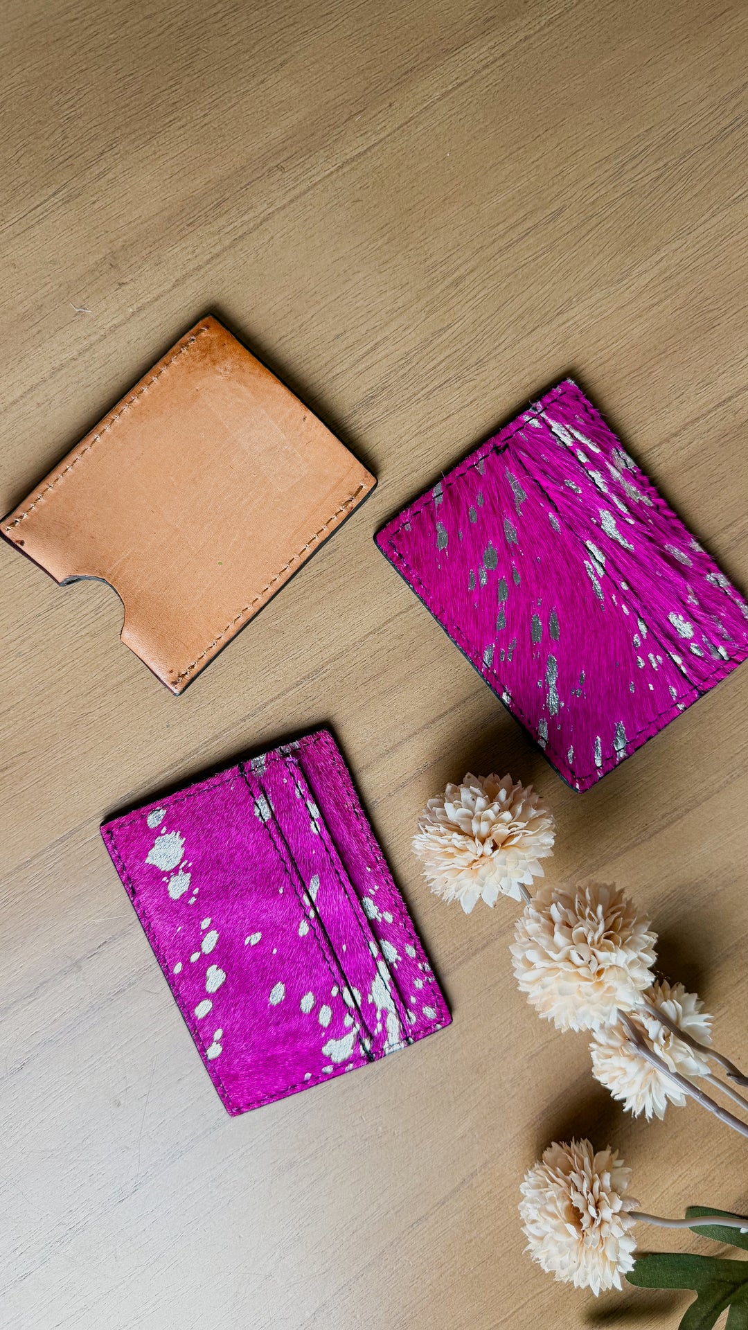 Three minimalist tooled leather cardholder wallets made of natural leather arranged on a light wood tabletop, alongside dried white flowers. One wallet is tan/caramel, and two are bright magenta hair on cowhide with sulver speckled patterns.