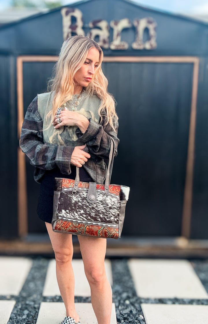A woman stands outdoors holding a large right hand concealed carry tote bag with a black-and-white genuine hair on cowhide and embossed leather green and burnt orange patterned trim. She wears a plaid shirt and black shorts in front of a dark building with a sign that reads Barn.