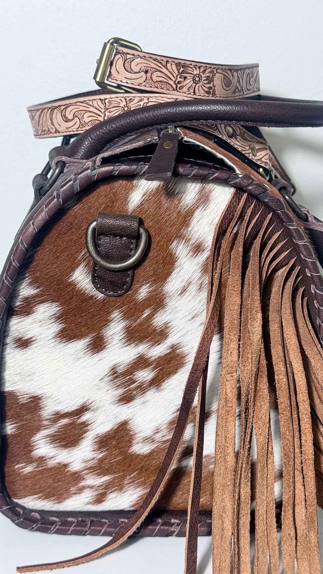 Side of cowhide Western satchel showing long brown leather fringe, whipstitch edging, strap attachment ring and detachable floral tooled strap.