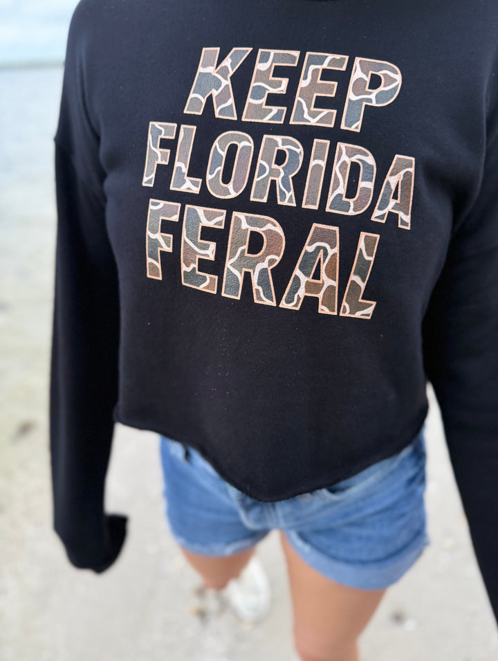 Woman wearing a black long-sleeve cropped hoodie with 'KEEP FLORIDA FERAL' camo text on a beach.