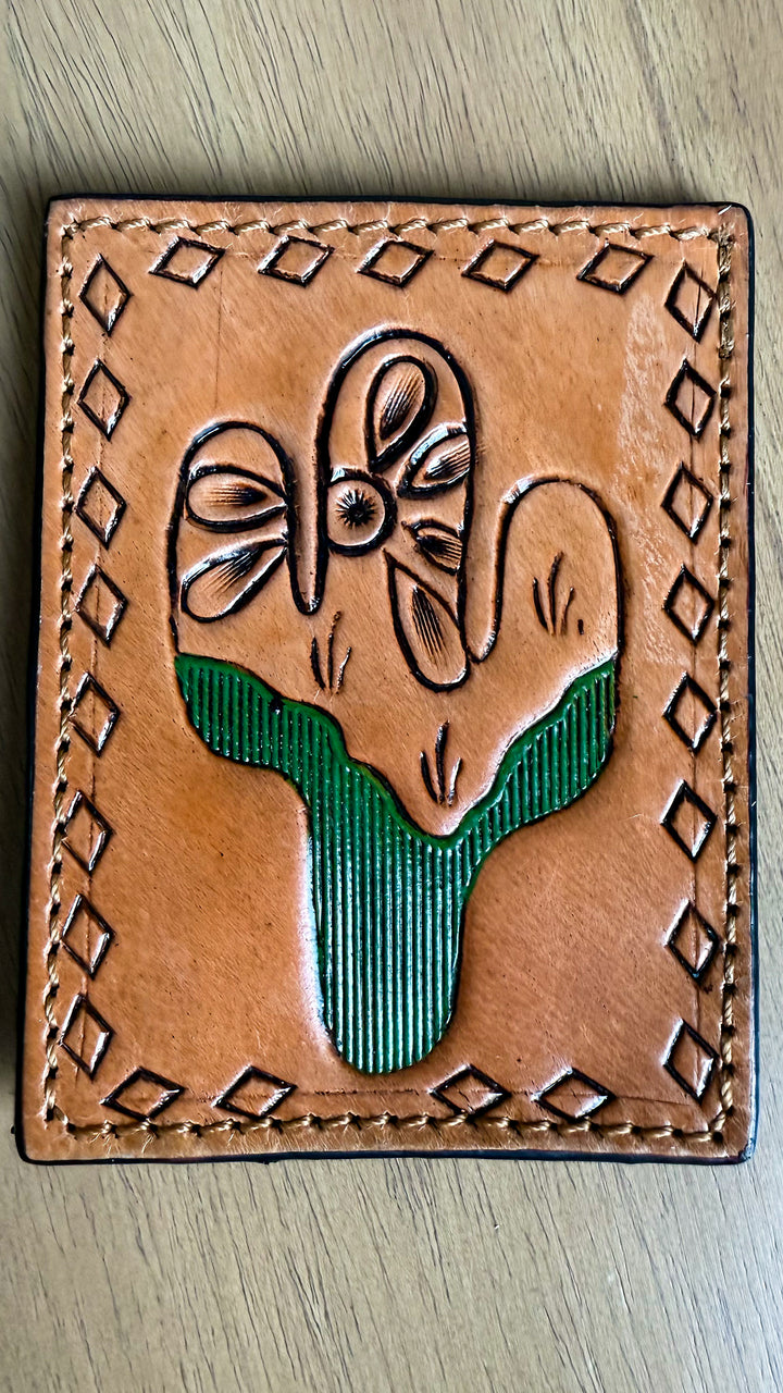 A square tan leather wallet or card holder featuring a tooled cactus design with a green striped center, photographed on a light wood surface.