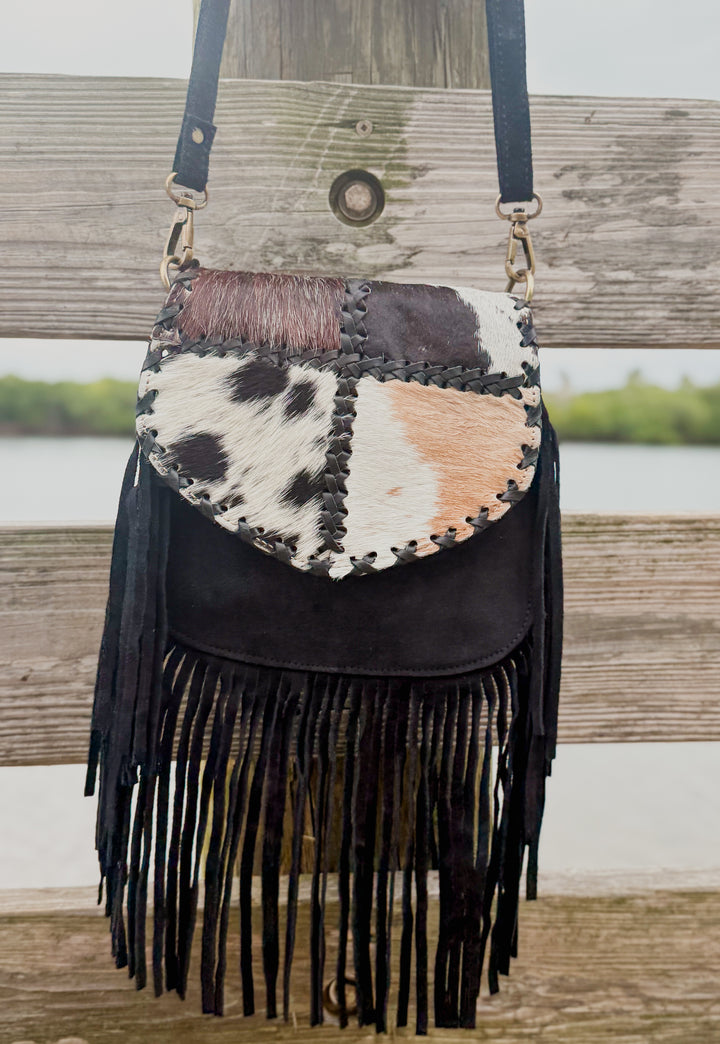 Black suede fringe crossbody purse with patchwork cowhide flap hanging on wooden railing
