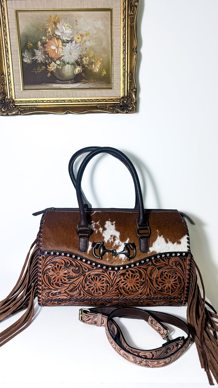 The Olde Money structured Western satchel with brown and white cowhide, floral tooled leather, horsebit accent, rhinestone studs and fringe.