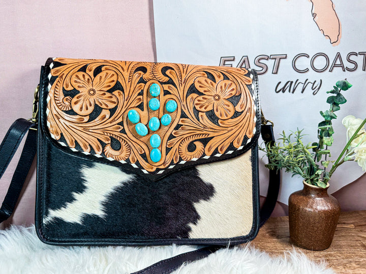 Cowhide leather handbag with floral tooled design and turquoise stones on a wooden surface.