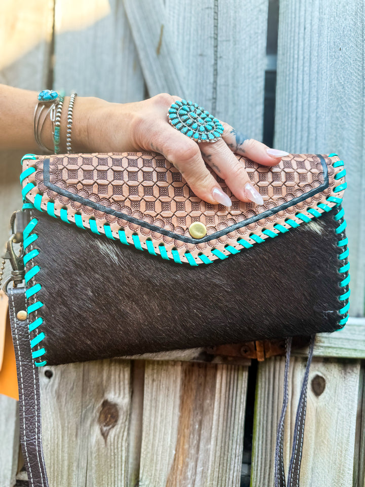 Hand holding a brown leather cowhide clutch with turquoise accents against a wooden fence background