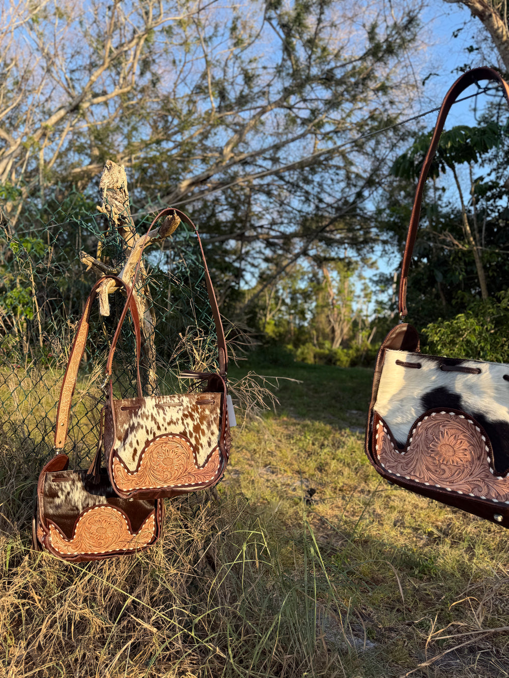 Western cowhide bucket bag with hand-tooled leather detailing and adjustable strap, shown outdoors with natural rustic background