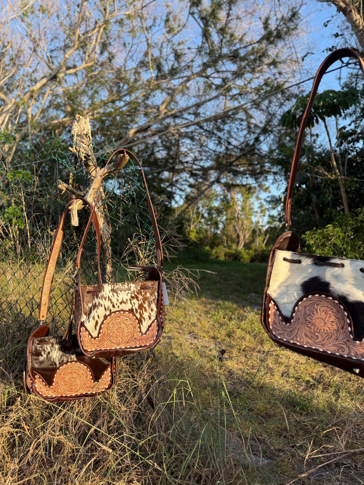 Western cowhide bucket bag with hand-tooled leather detailing and adjustable strap, shown outdoors with natural rustic background