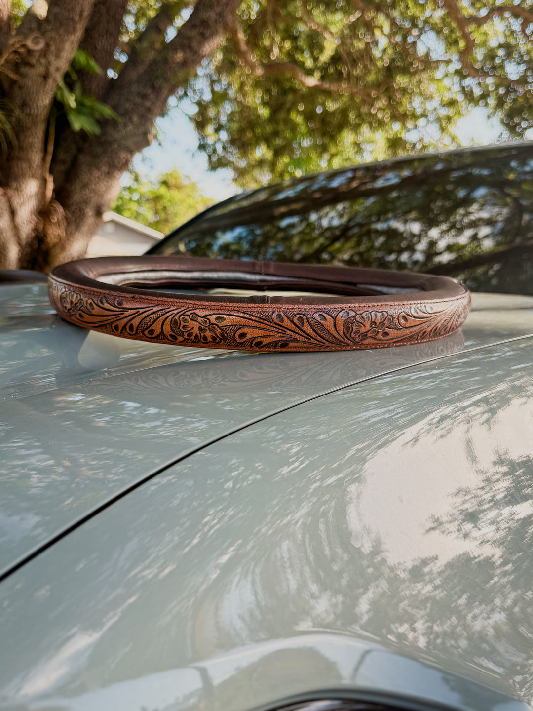 Coffee brown genuine leather steering wheel cover with western floral tooling detail displayed on a vehicle hood outdoors under oak trees.