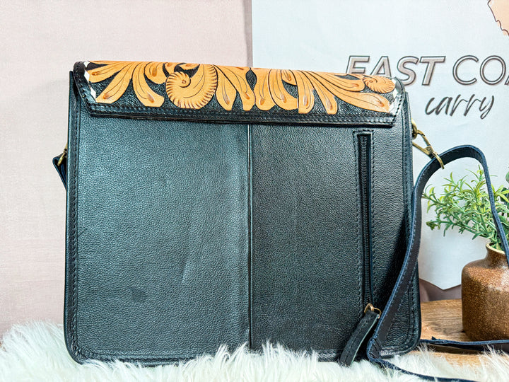 Black handbag with floral design on flap, showing exterior right zipper pocket for discreet carry placed on a white surface with a blurred background.