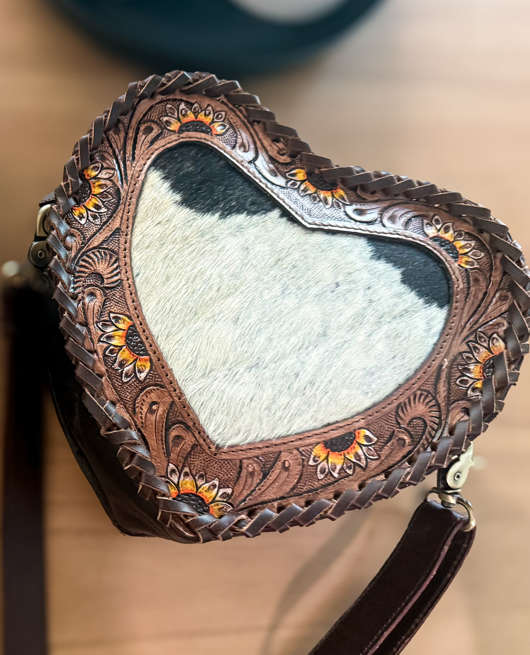 A heart-shaped leather crossbody bag with intricate floral tooling, braided leather trim, and a cowhide center, shown on a wooden surface.