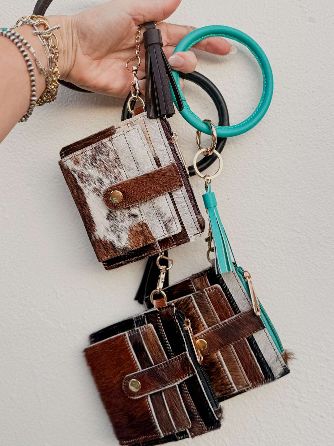 Cowhide leather bangle wristlet wallets with tassels and assorted hide patterns held in hand.
