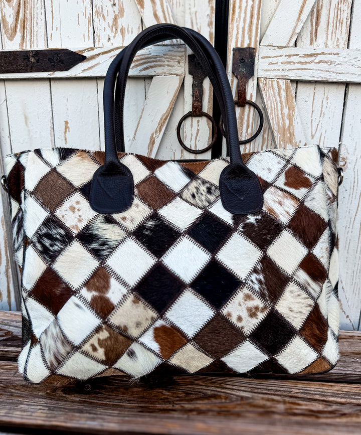 Checkered patterned cowhide western tote bag with rich brown handles on a wooden surface with detachable strap included 