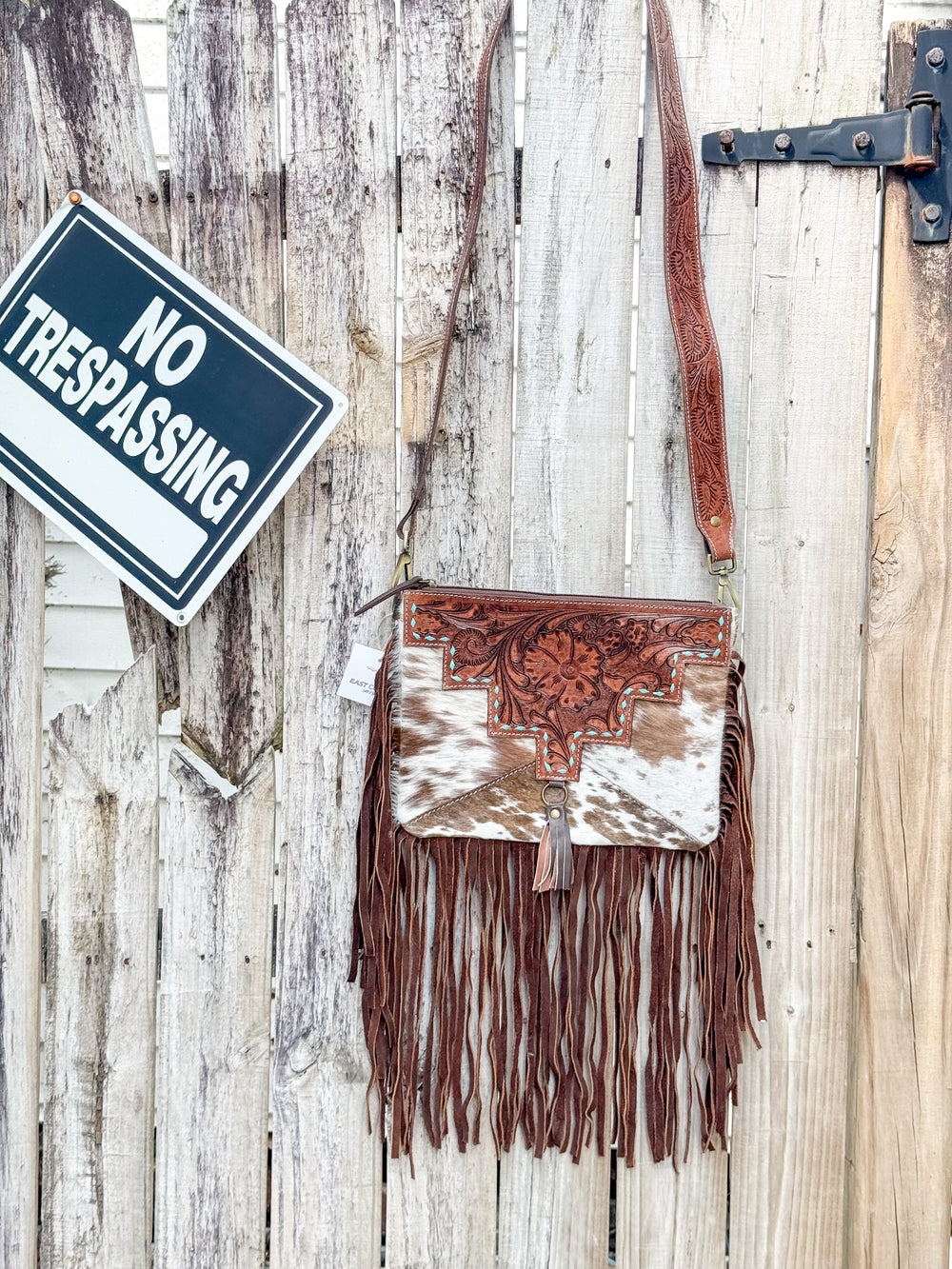 A brown and white cowhide crossbody bag with turquoise stitching and long brown leather fringe hanging against a wooden background with a 'No Trespassing' sign.