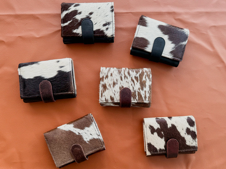Six small trifold women’s wallets with natural hair on cowhide-patterned exteriors and dark brown or black leather snap closures and accents , arranged on a textured orange surface.