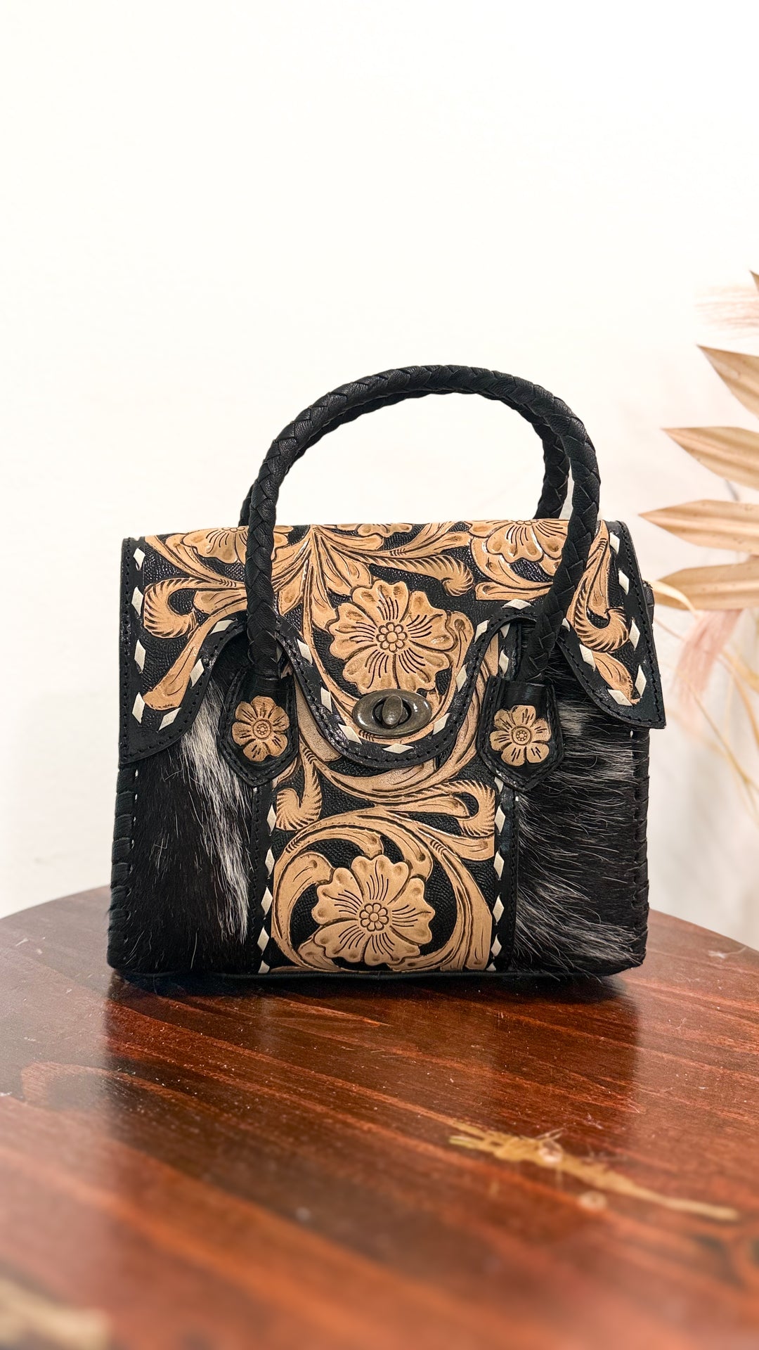 The Ranch Wife Carryall Mini black and white cowhide purse with natural floral tooled leather flap, antique bronze turn-lock closure and braided black leather handles.