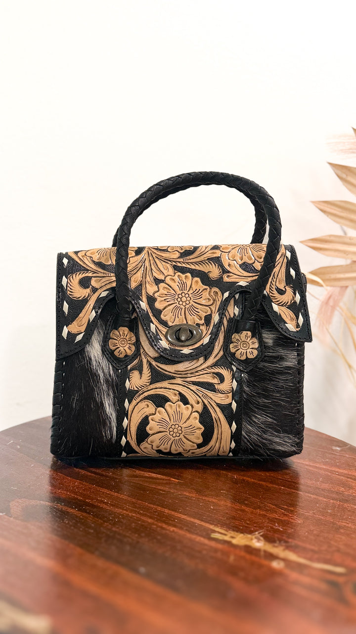 The Ranch Wife Carryall Mini black and white cowhide purse with natural floral tooled leather flap, antique bronze turn-lock closure and braided black leather handles.