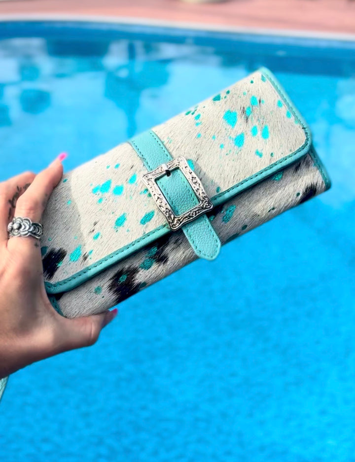 Hand holding a black and white speckled cowhide wallet with turquoise leather accents and silver buckle against a blue pool background