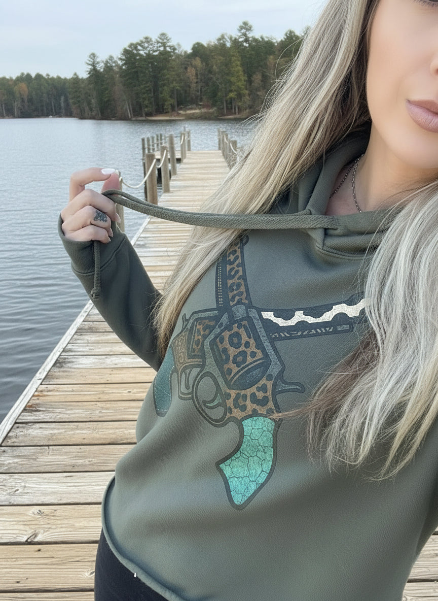Model wearing olive green cropped hoodie with turquoise and leopard crossed pistol design standing on wooden dock by lake — Western coastal style.