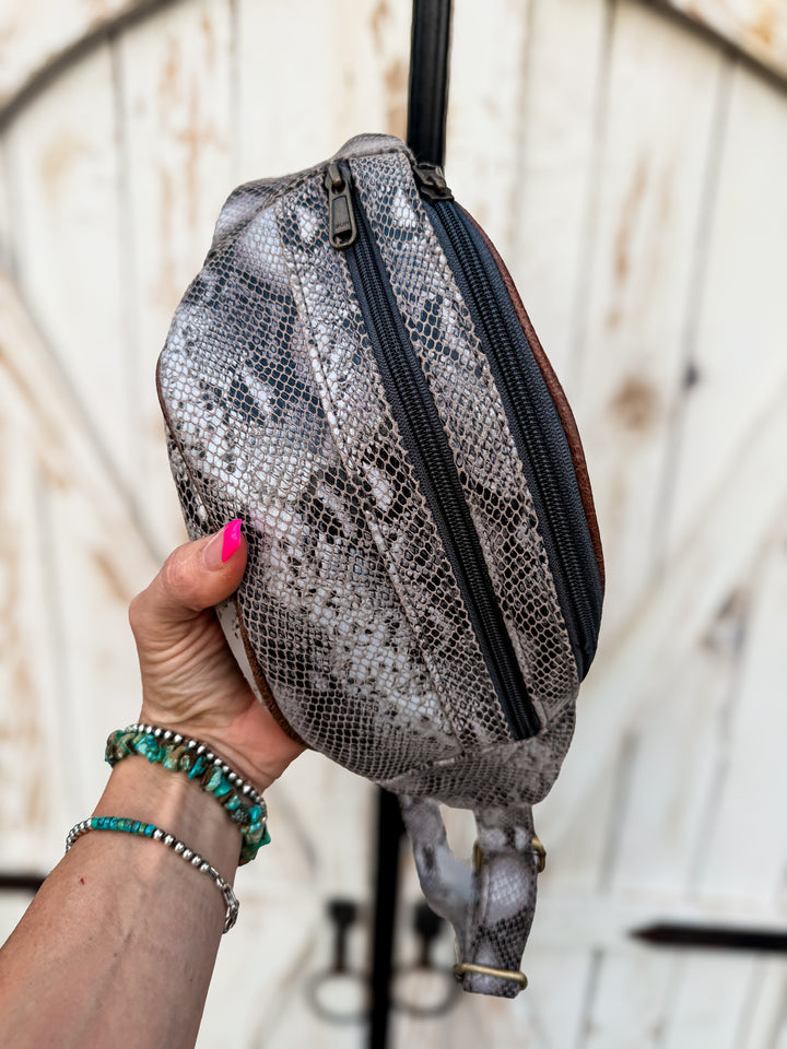 Hand holding a Grey genuine leather snake print belt bag with adjustable strap and dual zipper compartments.