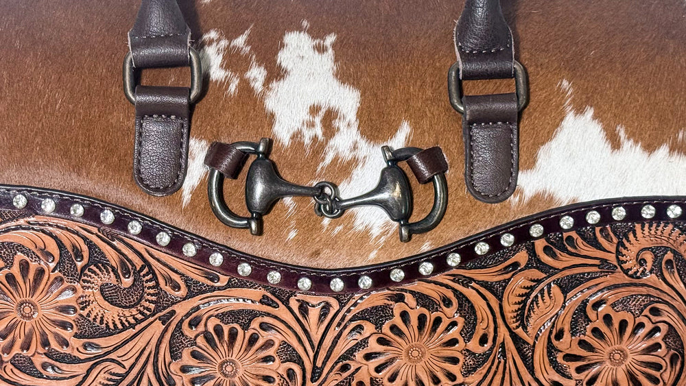 Close-up of dark metal horsebit accent on brown and white cowhide purse with rhinestone stud trim and floral tooled leather.