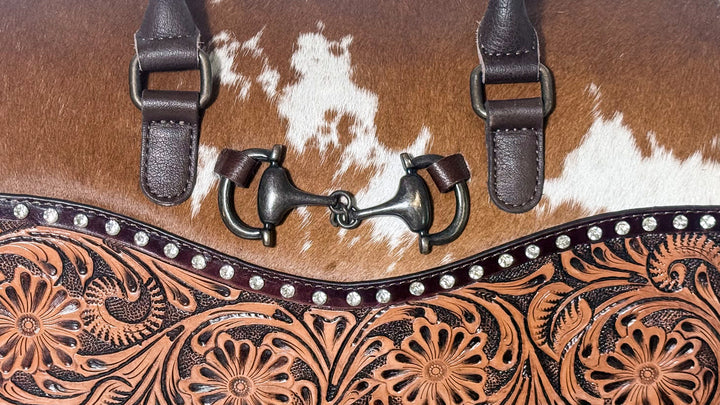 Close-up of dark metal horsebit accent on brown and white cowhide purse with rhinestone stud trim and floral tooled leather.