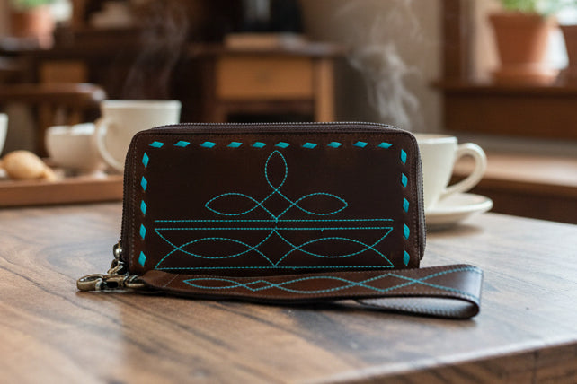 A dark brown leather wristlet clutch wallet with turquoise blue boot stitch design embroidery, resting on a wooden table with a blurred background of steaming coffee cups.