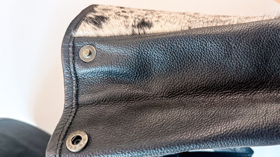 Close-up of expandable side panel on black leather backpack showing metal snap fasteners and textured leather construction.