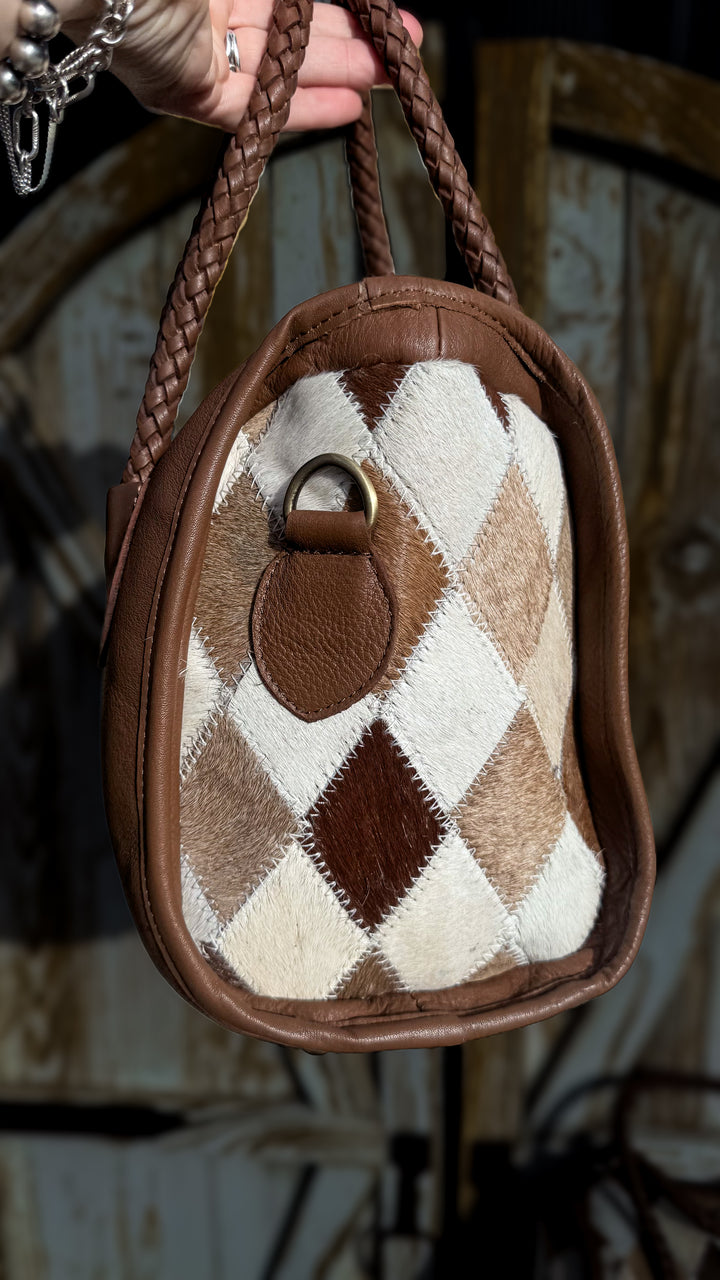 Side view of Soft & Strong cowhide satchel showing diamond patchwork hair-on hide, brown leather trim and metal strap attachment.