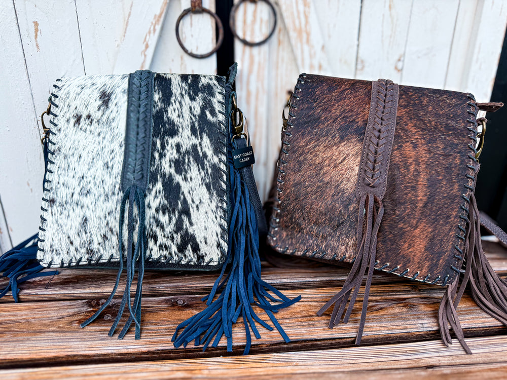Two western-style fringe concealed carry handbags displayed side by side on a wooden surface. The left bag features a black and white cowhide pattern with dark blue fringe and strap, and the right bag is made of distressed brown leather with a matching brown strap and fringe.