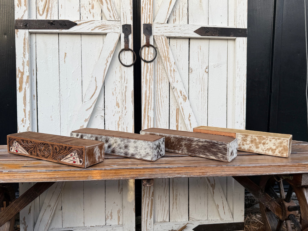 Four rectangular decorative western ring holder displays with rustic genuine hair on cowhide and carved leather patterns, displayed on a wooden table in front of distressed white barn-style doors.
