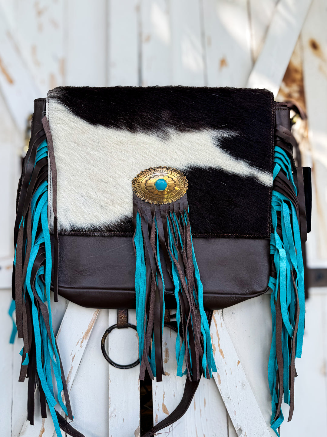 A western-style shoulder bag featuring a black and white cowhide flap, dark brown leather trim, and long turquoise fringe tassels, with a gold-tone concho buckle on the front.