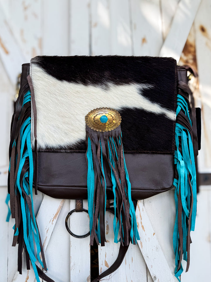 A western-style shoulder bag featuring a black and white cowhide flap, dark brown leather trim, and long turquoise fringe tassels, with a gold-tone concho buckle on the front.